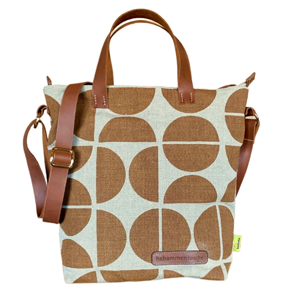 Die Hebammentasche "Pop" in ocker ist mit kurzen und langen Lederhenkeln ausgestattet. Die Vorderseite ist mit einer Lederaufschrift "hebammentasche" und einem halbkreisartigen Muster verziert.